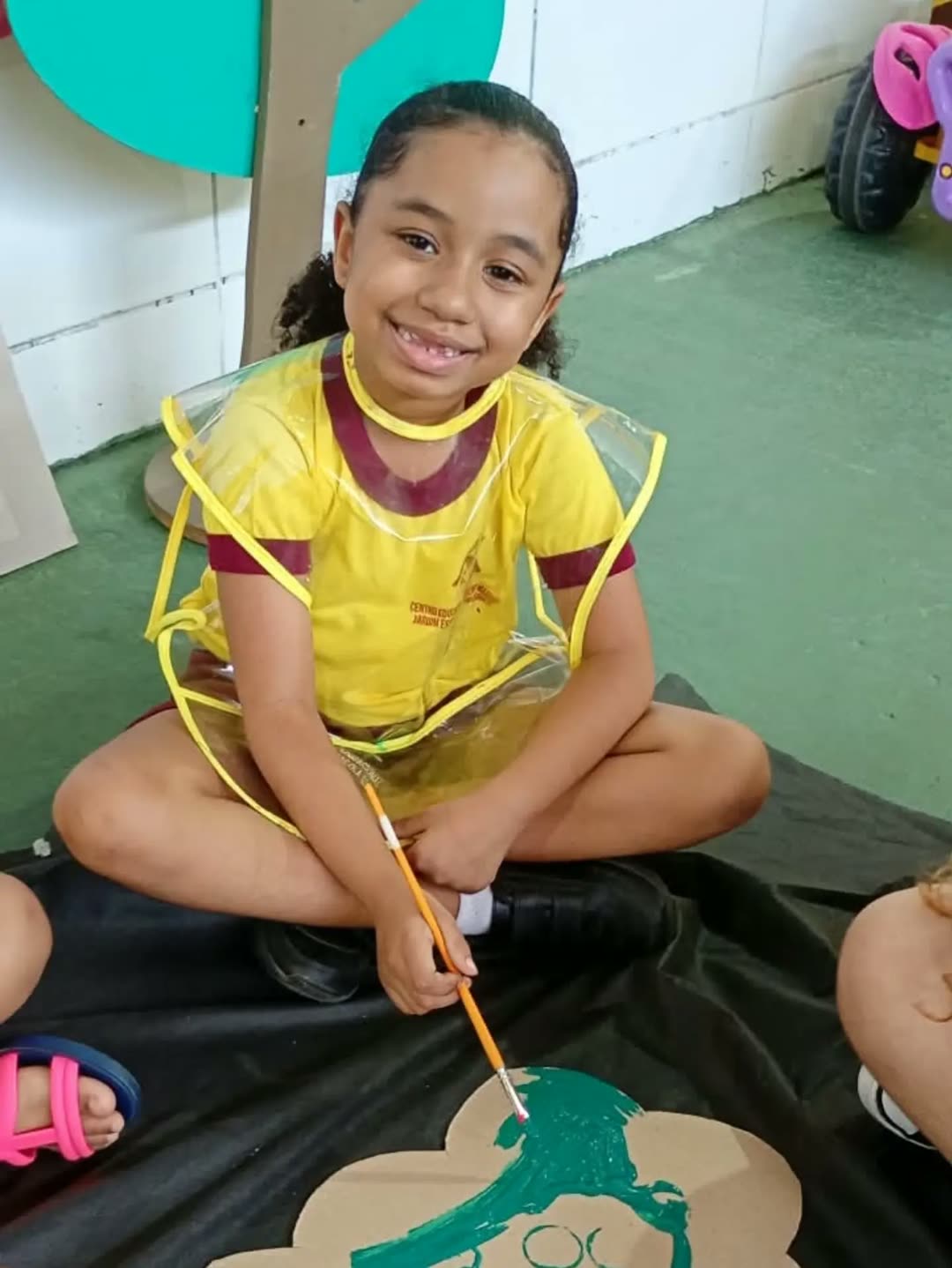 Menina sorrindo sentada no chão, com pincel na mão, pintando de verde a copa de uma árvore de papelão