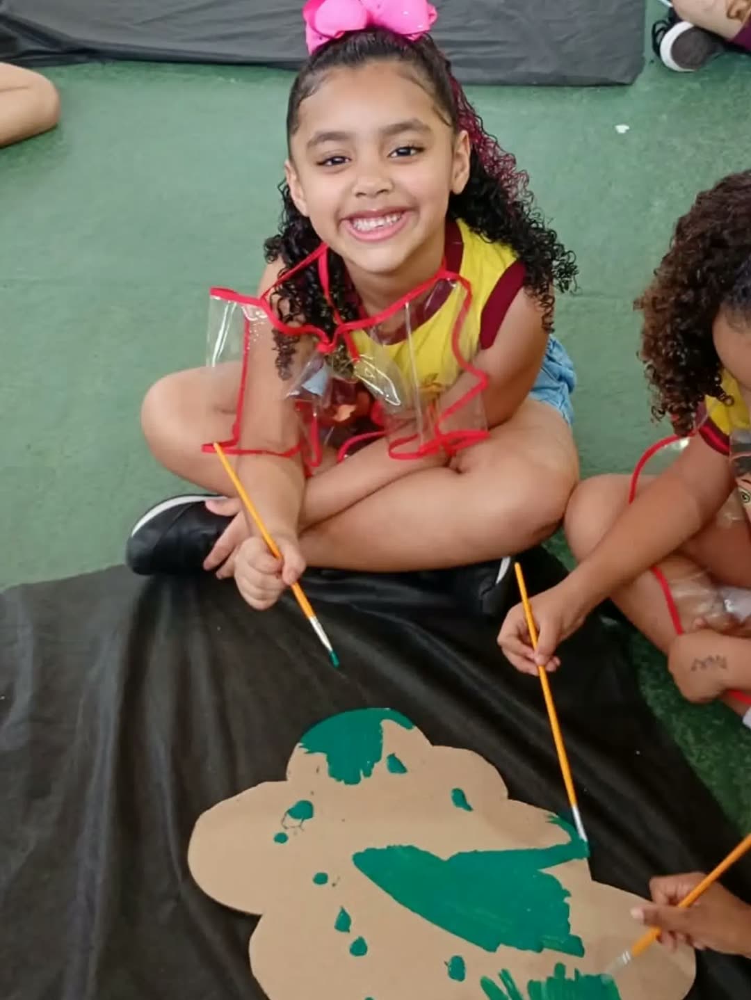 Menina com laço rosa no cabelo sorrindo enquanto pinta com a colega